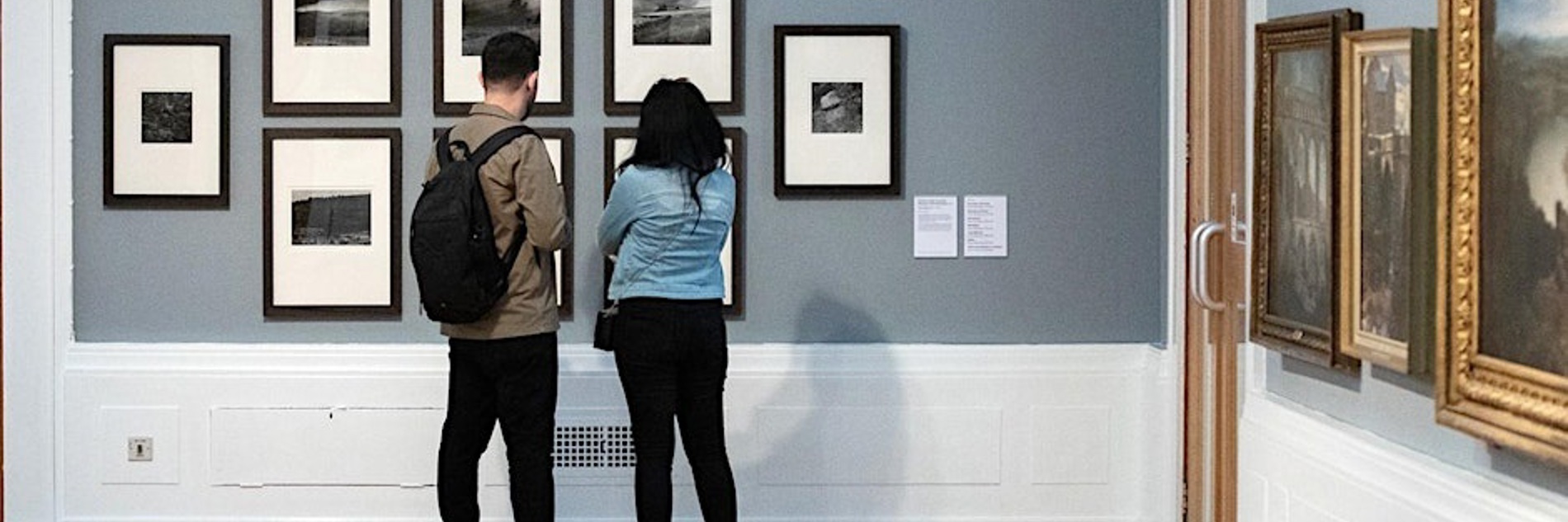 A photograph of two adults looking at a wall of black and white photographs in a gallery setting.
