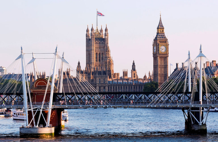 A photograph of the Hungerford Bridge in London, with the River Thames visible underneath and Big Ben and The Houses of Parliament visible in the background.