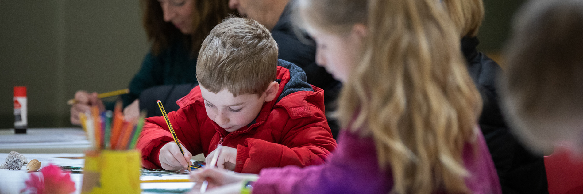 A photograph of a young boy drawing at a table. He is very focussed and there are other people sat at the table next to him, but they are out of focus. Paper and pots of pens are visible on the table in front of him