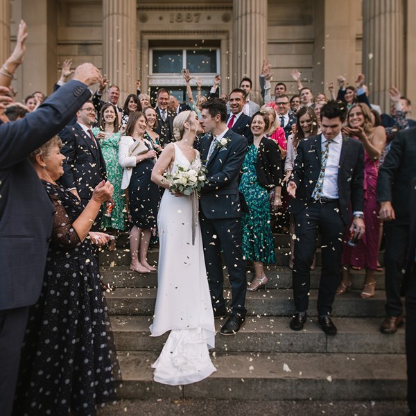 A bride in a wedding dress and a groom  in a suit kiss on teh steps of a historic building as wedding guests around them throw confetti. 