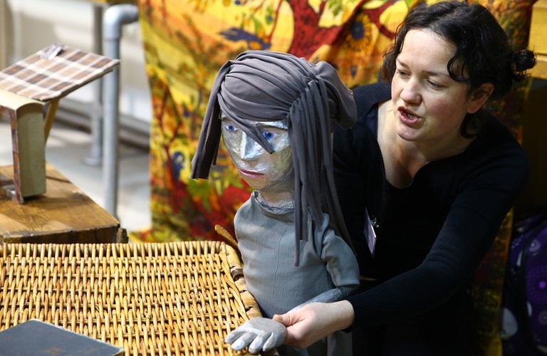 A person with a lifesized puppet, it is dressed in grey with grey fabric as hair, a small model of a building is on box behind them. 