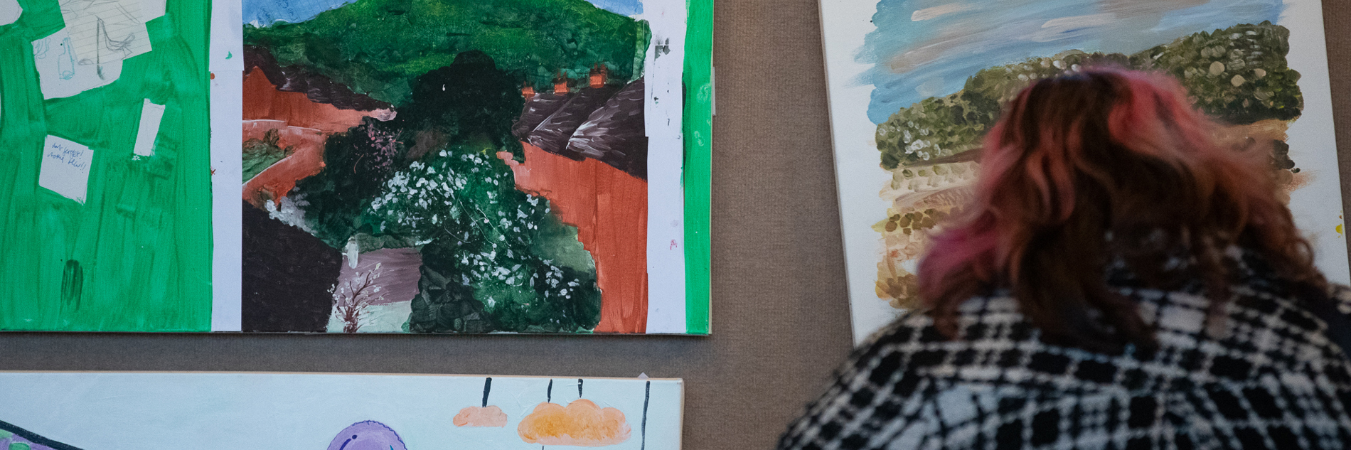 A person with shoulder length pink and black hair and a black and white check shirt in front of a display of three landscape artworks.