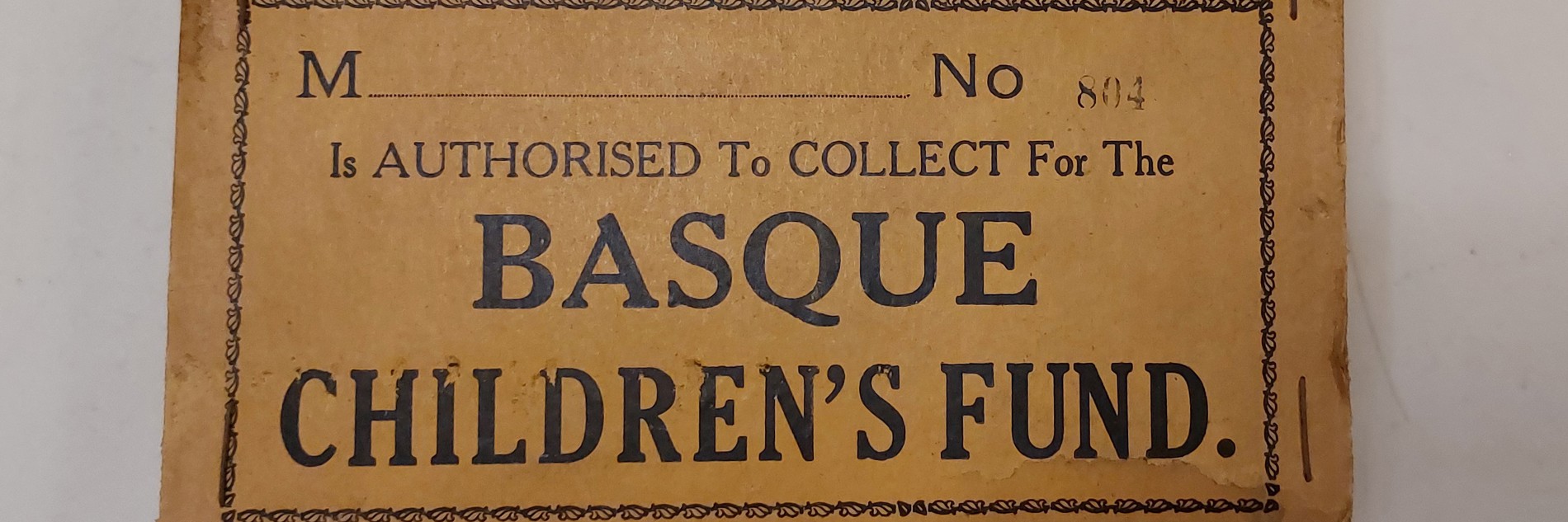 A photograph of a brown label. In black text, it reads: M No 84 is authorised to collect for the Basque children's fund. It is photographed against a white background. 
