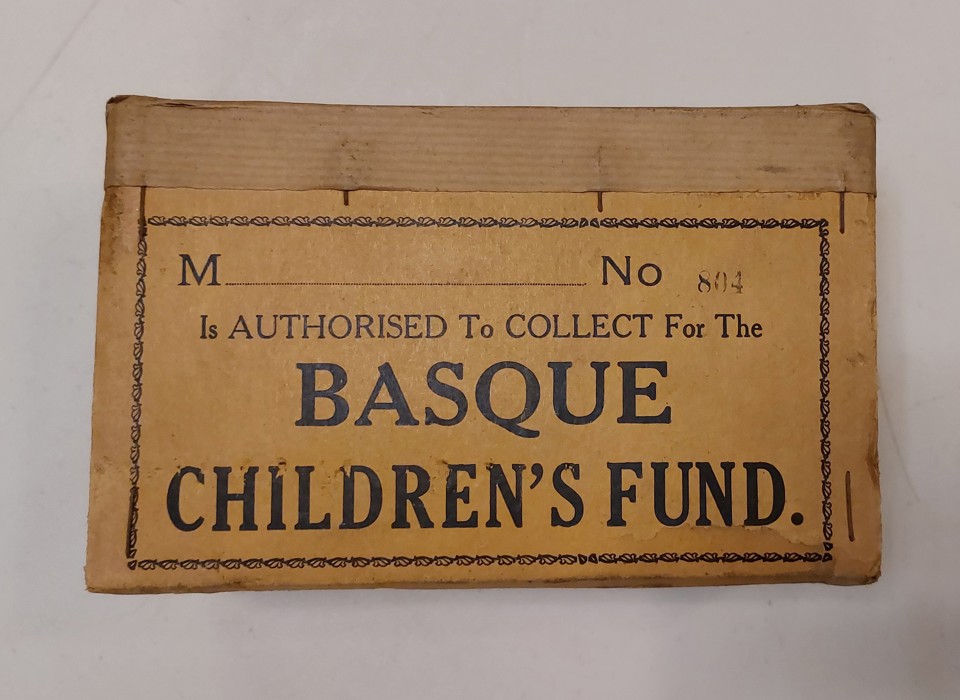 A photograph of a brown label. In black text, it reads: M No 84 is authorised to collect for the Basque children's fund. It is photographed against a white background. 
