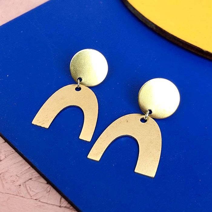 A pair of gold coloured, arch shaped earrings on a vivid clue and yellow background.  