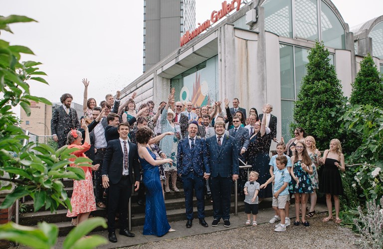 Large group of smiling, smartly dressed adults showering two adults wearing wedding outfits with confetti outside the Millennium Gallery. 