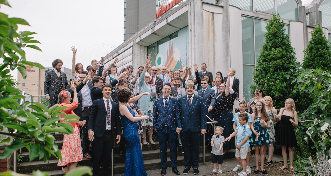 Large group of smiling, smartly dressed adults showering two adults wearing wedding outfits with confetti outside the Millennium Gallery. 