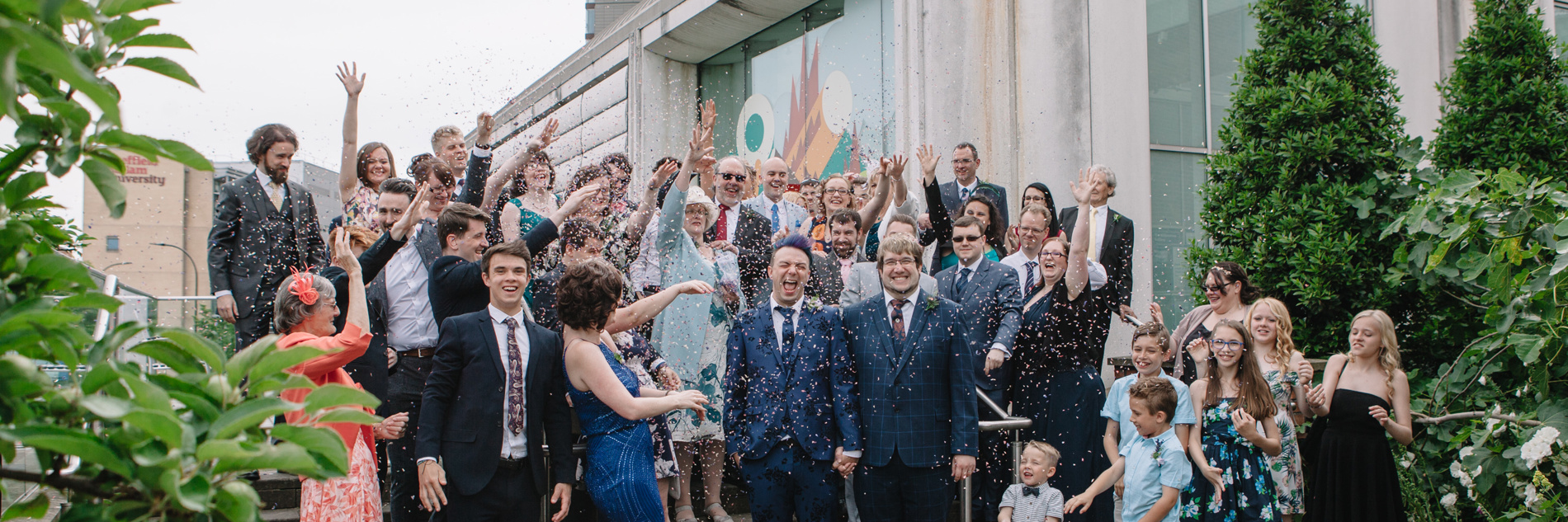 Large group of smiling, smartly dressed adults showering two adults wearing wedding outfits with confetti outside the Millennium Gallery. 