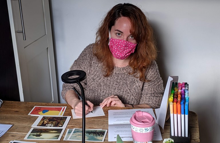 An adult in a facemask about to start writing, with a set of coloured pens to one side.