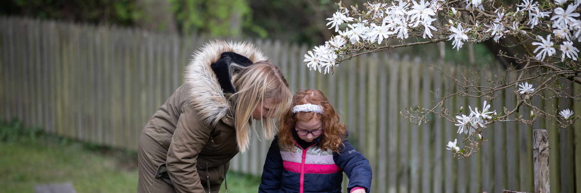 A photograph of a woman and a young girl looking at details in Abbeydale Industrial Hamlet. There a green leaves going up the wall and a tree full of white flowers to their right.