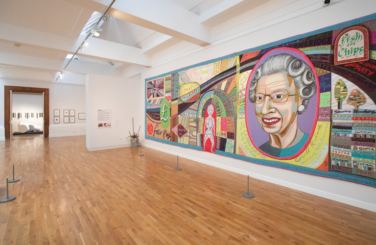 A photograph showing Grayson Perry's Comfort Blanket in Graves Gallery. It is a large patchwork quilt, with a large portrait of Queen Elizabeth 2nd on it.