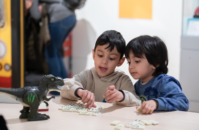 A photograph of two children playing with dominoes. On the table next to them is a toy dinosaur. 
