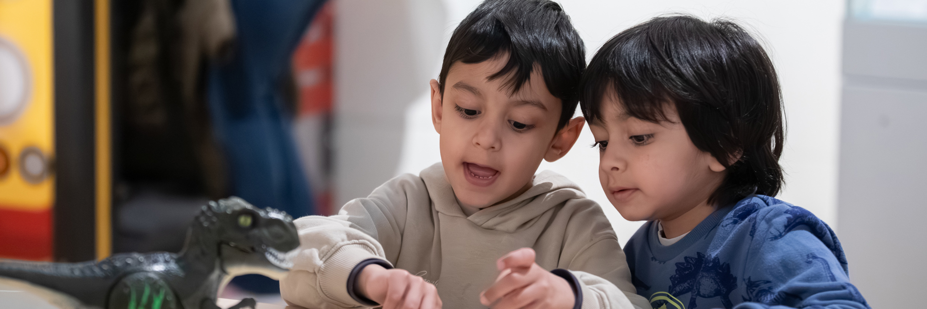 A photograph of two children playing with dominoes. On the table next to them is a toy dinosaur. 