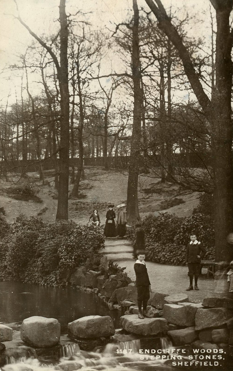 A black and white historic photograph showing a young person stood on stepping stones across a river in a park. Another young person is stood on the footpath to their right, while two people in dresses stand on further kalong the path.