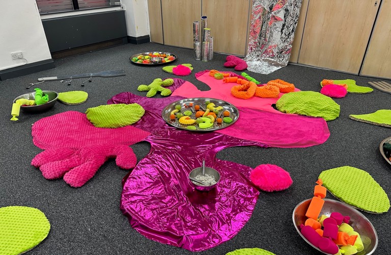 A photograph of an indoor space set up for toddler play, with lots of soft cushions, shiny fabric, and sensory toys.