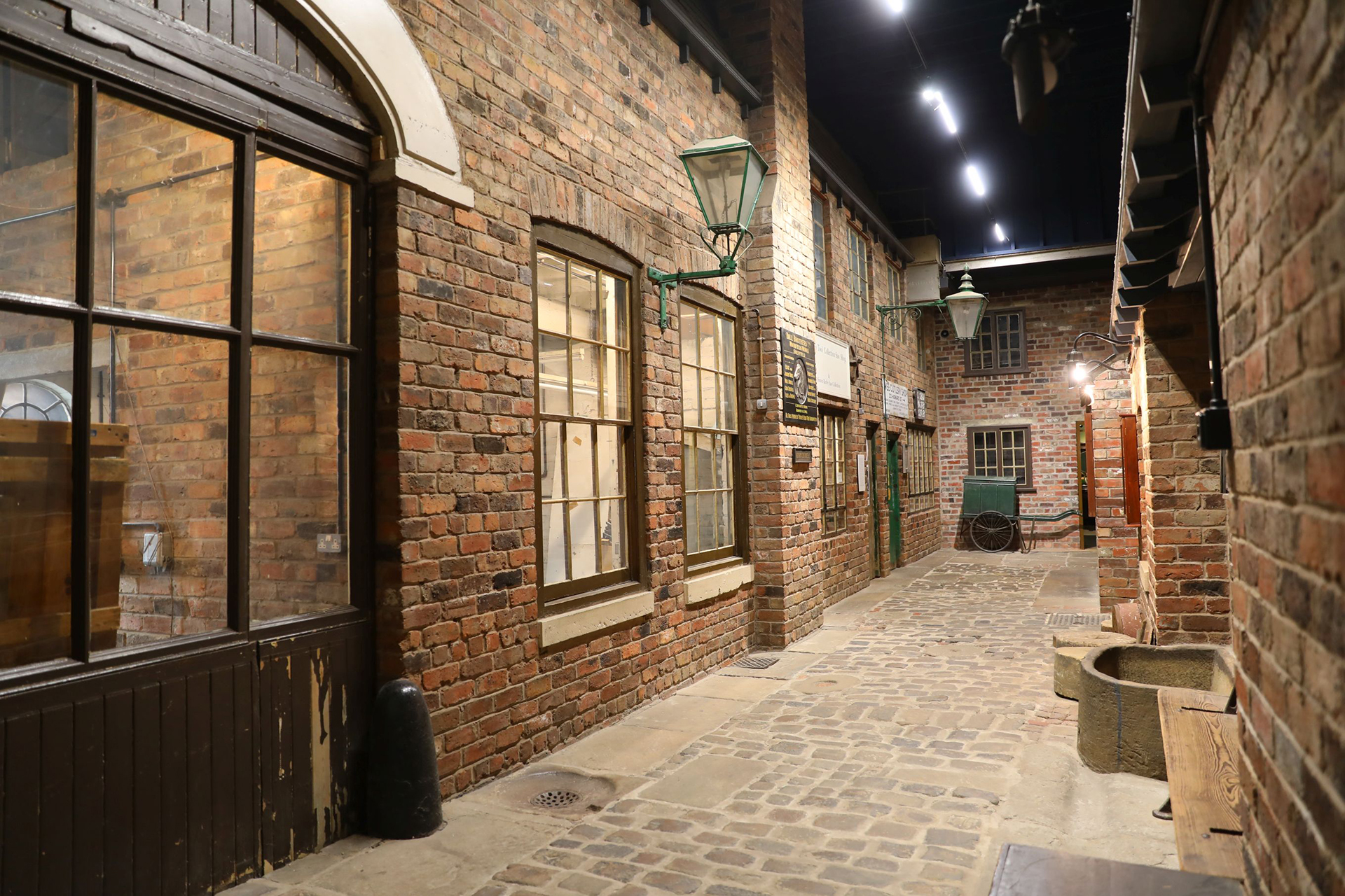 A perspective view along a restored cobbled street. The red brick walled workshops have multi-paned sash windows. There are two green wall mounted gas lamps and a green handcart at the end of the street.