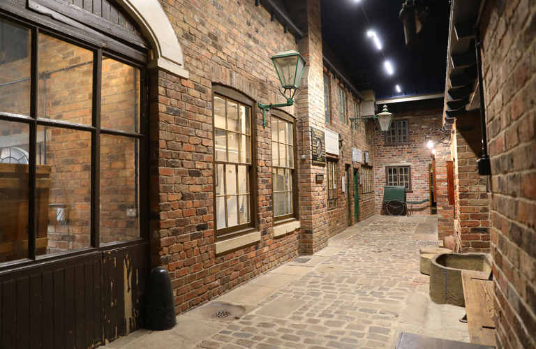 A perspective view along a restored cobbled street. The red brick walled workshops have multi-paned sash windows. There are two green wall mounted gas lamps and a green handcart at the end of the street.