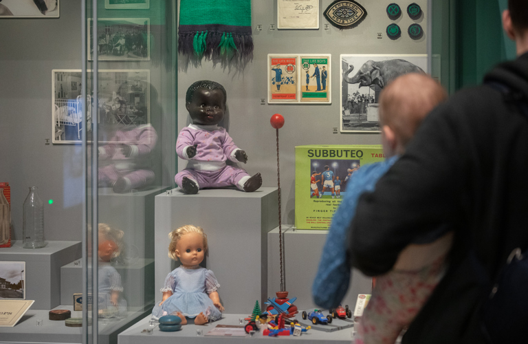 A photograph of a man holding a baby, looking at a glass display case of old childhood toys' baby dolls, subbuteo, a yoyo and some lego