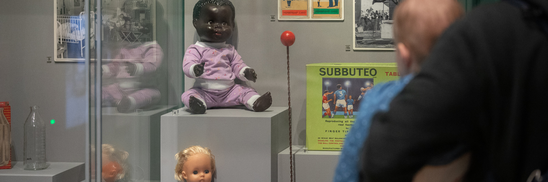 A photograph of a man holding a baby, looking at a glass display case of old childhood toys' baby dolls, subbuteo, a yoyo and some lego