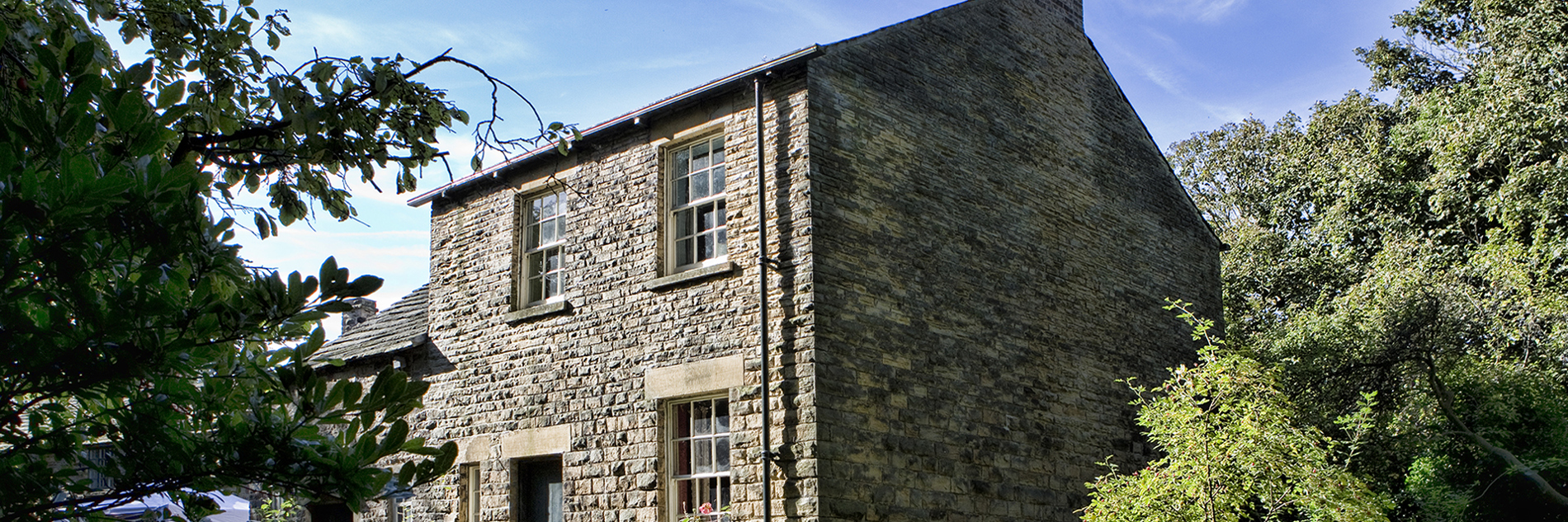 Outside of stone built cottage with Georgian windows and chimney stack in a wooded location in bright sunshine.