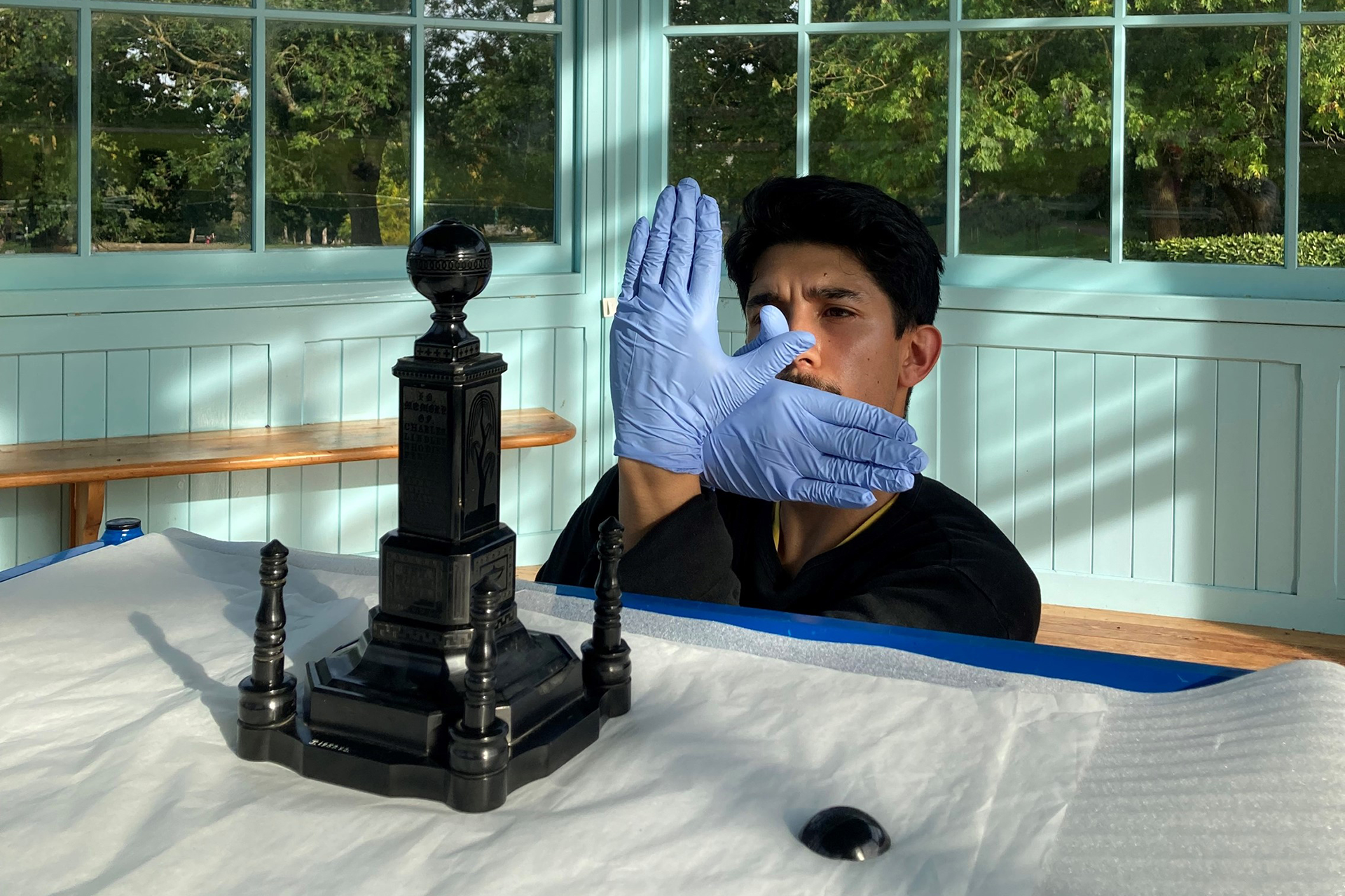 A person behind a table wearing blue gloves making an overlapping hand gesture in an L shape. On the table is a small version of a monument, with four columns surrounding a large central square tiered column topped with a sphere.