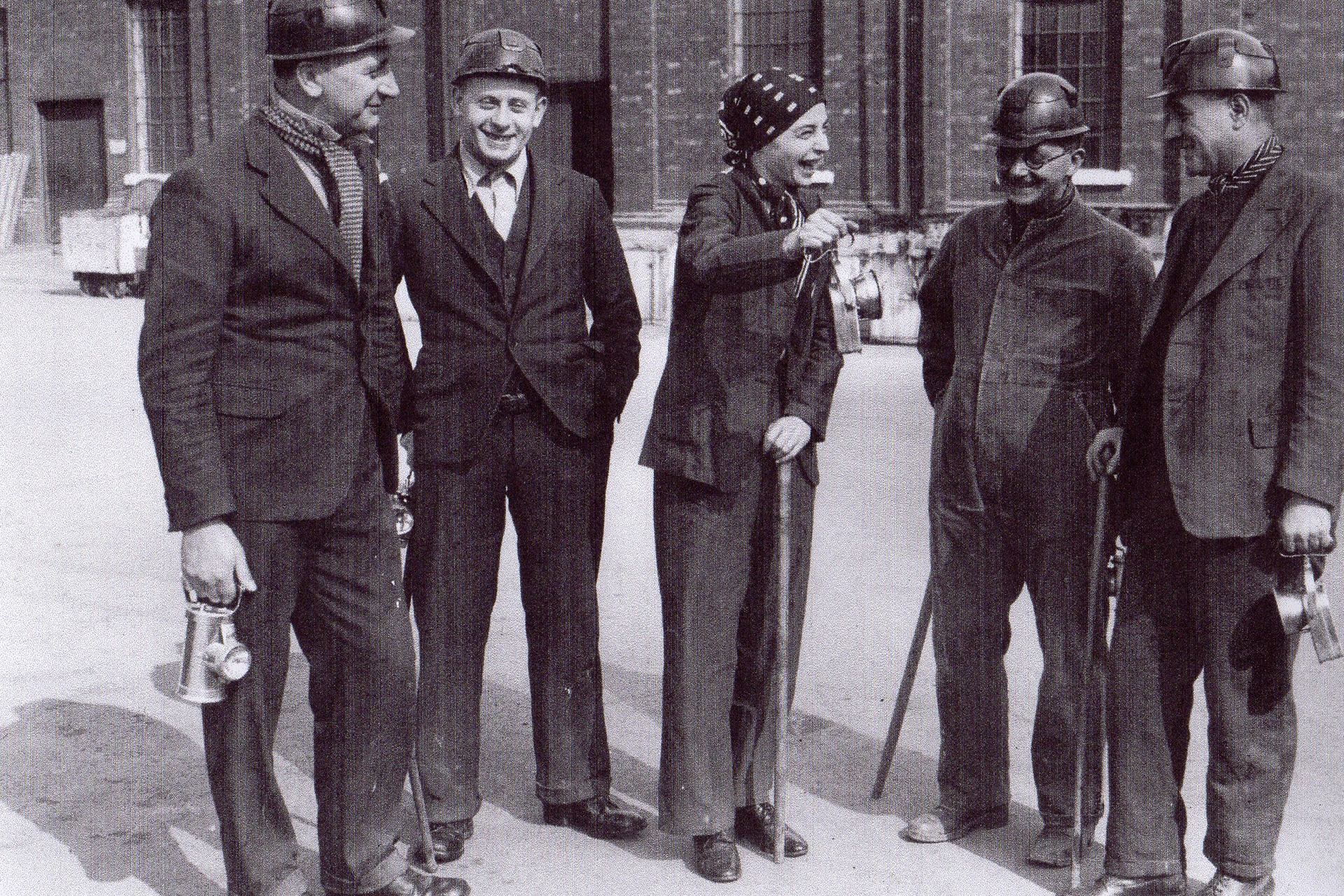 Black and white photograph of 5 people in suits standing outside in an industrial yard. Monica Maurice is in the middle of the group, wearing a trouser suit and a headscarf