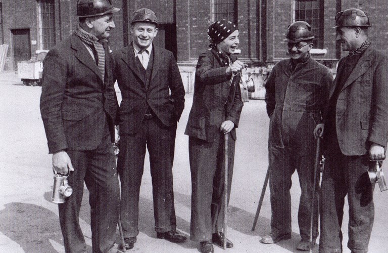 Black and white photograph of 5 people in suits standing outside in an industrial yard. Monica Maurice is in the middle of the group, wearing a trouser suit and a headscarf