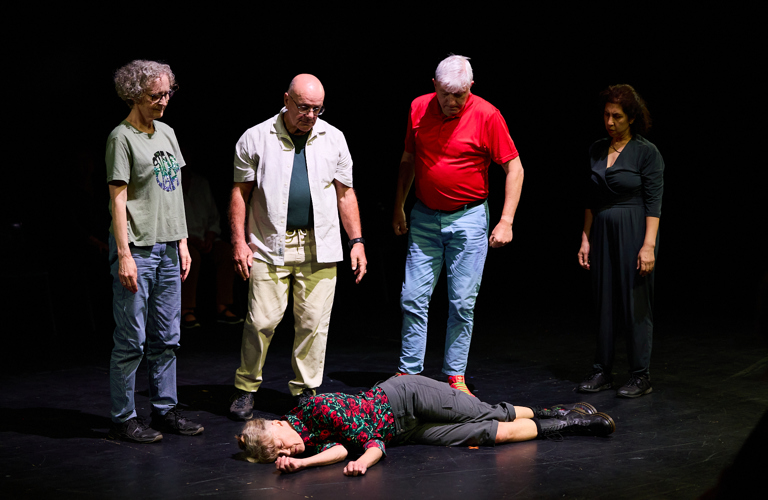 A photograph of a performance. Five performers are on a black stage. One lies on the floor, four people stand above, looking down at them. 