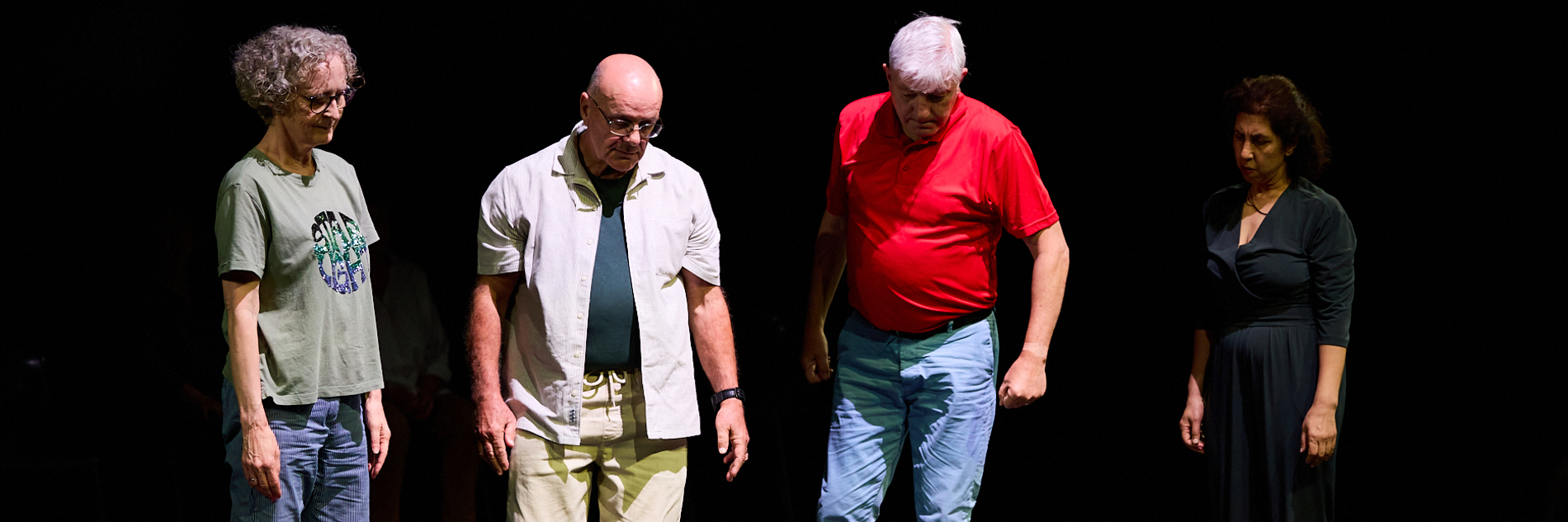 A photograph of a performance. Five performers are on a black stage. One lies on the floor, four people stand above, looking down at them. 