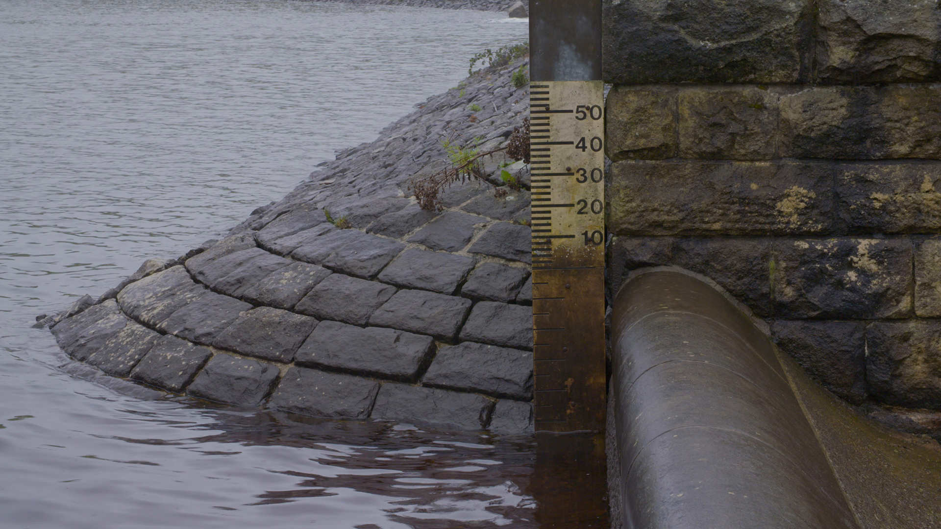 A photograph of an old stone brick sloping wall on the edges of a reservoir, with a measure to monitor the water level in the centre. 