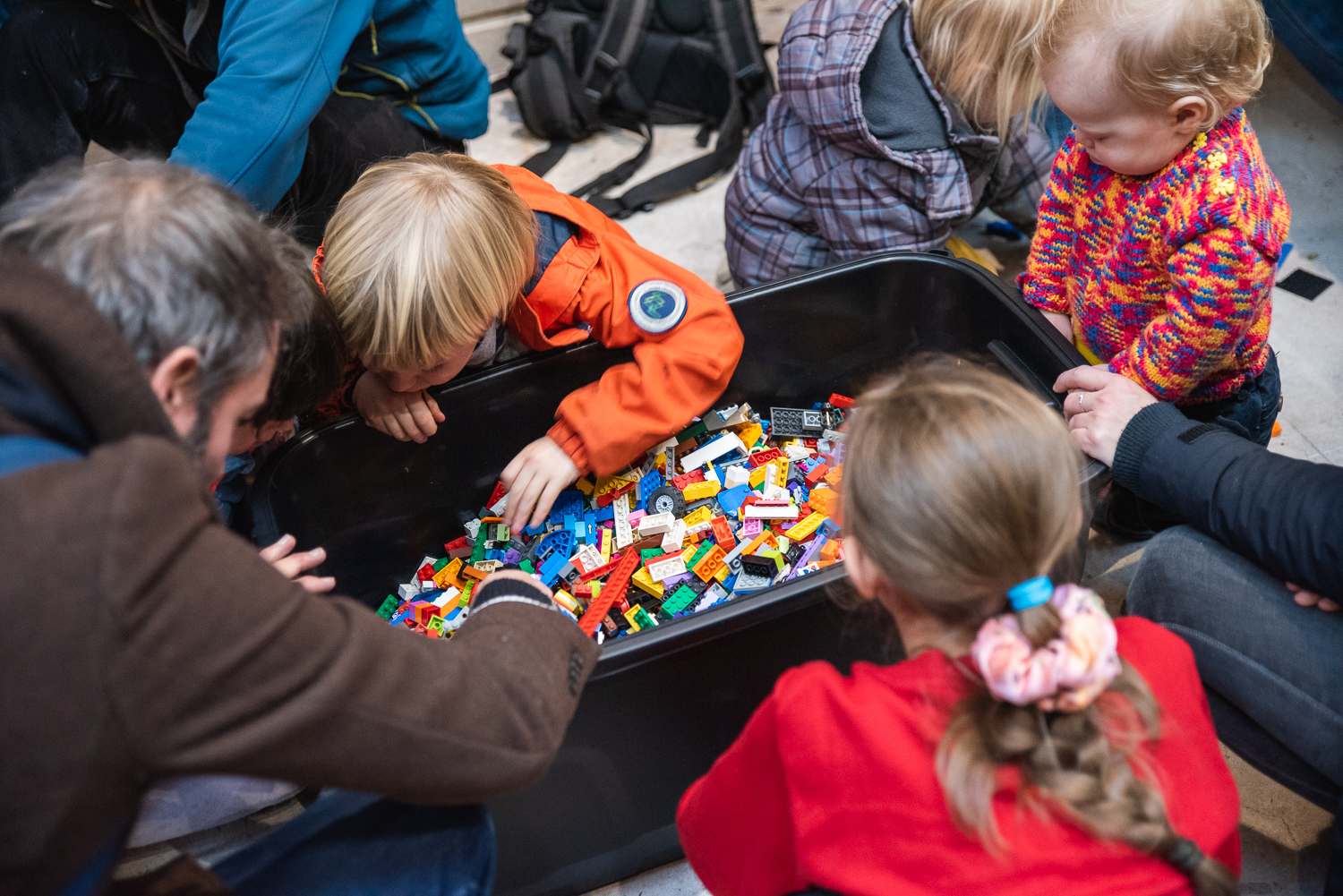 A group of children and adults kneel around a box of LEGO selecting bricks.