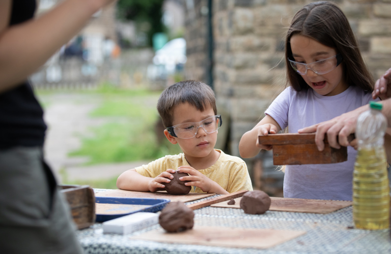 A photograph of two children with brown hair wearing goggles and rolling brown clay. One holds a wooden mold.