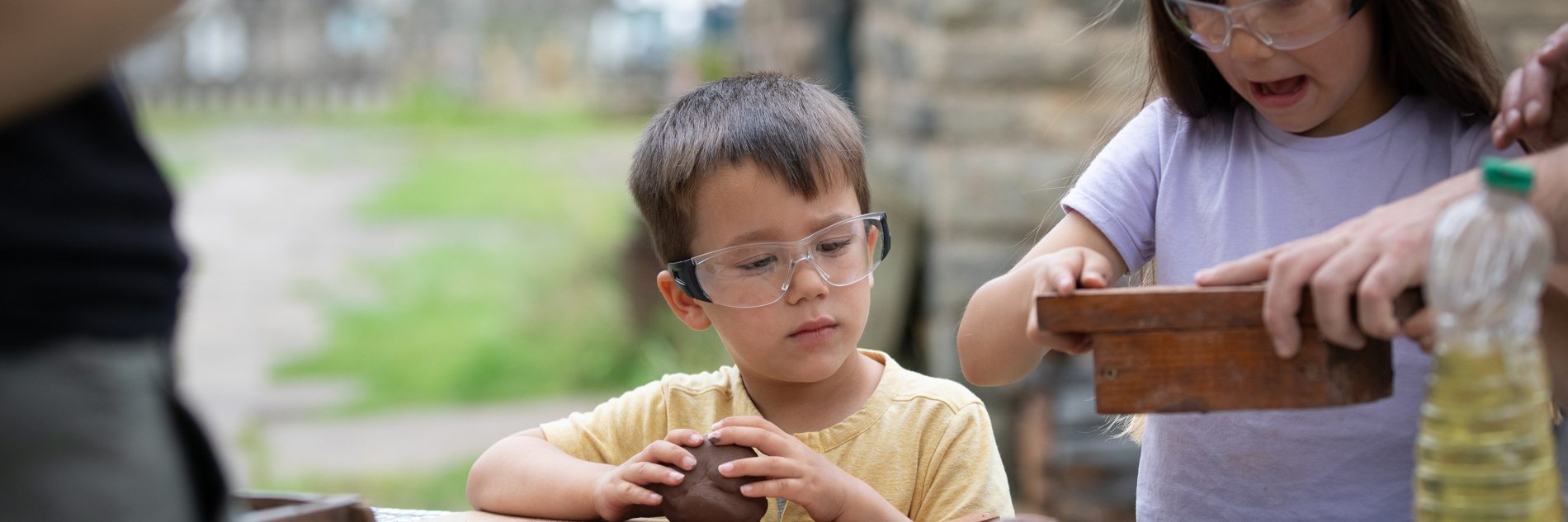 A photograph of two children with brown hair wearing goggles and rolling brown clay. One holds a wooden mold.