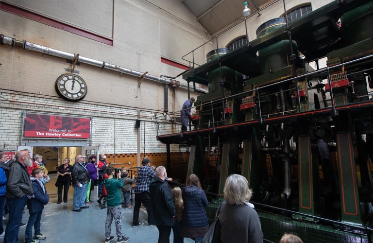 A group of visitors at Kelham Island Museum stand behind a metal barrier to watch an engineer run the River Don engine. The large steam engine is dark green, red, black and gold. The mechanics are all visible. It has exposed pistons, three cylinders at the top and two platforms around it.