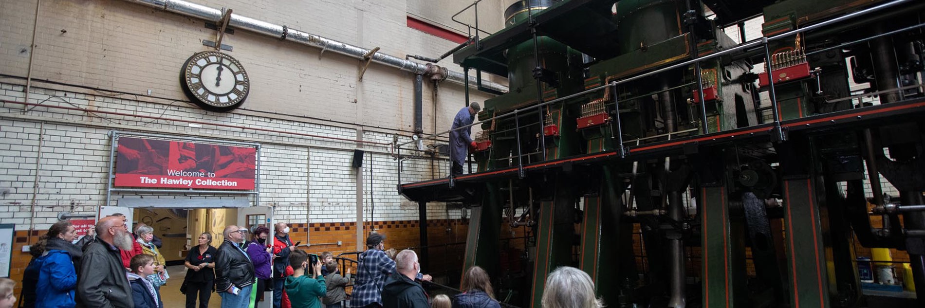 A group of visitors at Kelham Island Museum stand behind a metal barrier to watch an engineer run the River Don engine. The large steam engine is dark green, red, black and gold. The mechanics are all visible. It has exposed pistons, three cylinders at the top and two platforms around it.