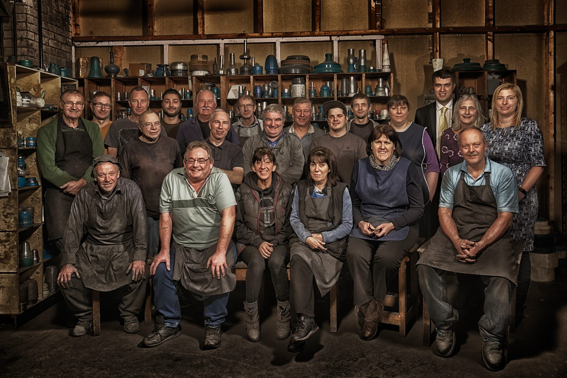 22 people pose in three rows in a workshop. The people in the front row are seated on stools and the majority of people featured are wearing aprons. Behind the people are shelves featuring various examples of part-made pewter objects and other items related to their production.
