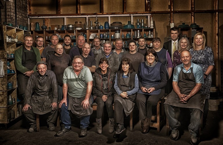 22 people pose in three rows in a workshop. The people in the front row are seated on stools and the majority of people featured are wearing aprons. Behind the people are shelves featuring various examples of part-made pewter objects and other items related to their production.