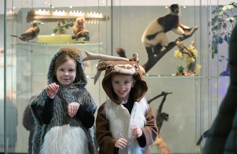 Two young children are dressed in animal costumes, standing in front of glass cabinets with taxidermied animals inside. 