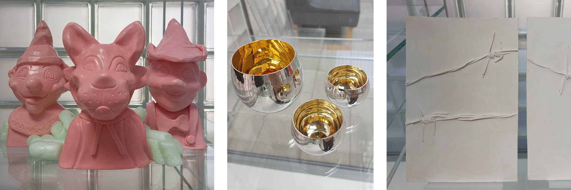 A composite of three images. Images (left to right): An image of three pink sculptural works presented in a glass display cabinet. On the right there is a clown, middle is an animal wolf and on the left is an interpretation of Pinocchio, all three are busts. There are styrofoam packing peanuts inbetween the pieces.  Then a photograph of one metal bowl and two smaller whiskey shots in a glass display case, silver on the outside and gold on the inside, in two different sizes. Finally, a photo of two white canvases in a glass display cabinet. The left piece has two pieces of barbed wire across the canvas, with paint covering them. The right piece has one piece of barbed wire, also painted over.