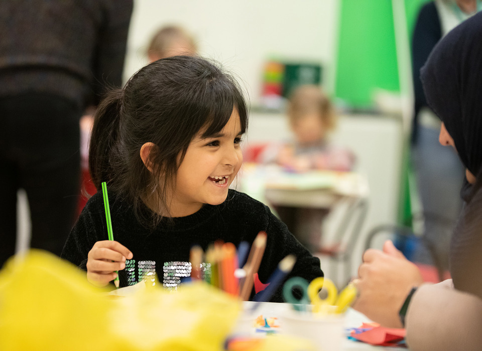 A young child is sitting at a table and is smiling and holding a green pencil. In the foreground is some yellow tissue paper and pots of scissors and pencils.