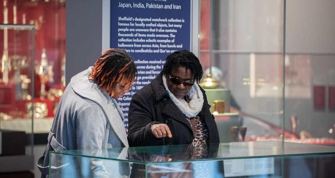 Two adults are looking at a glass cabinet in the gallery, one of the adults is pointing. There is a dark blue gallery sign behind them. 