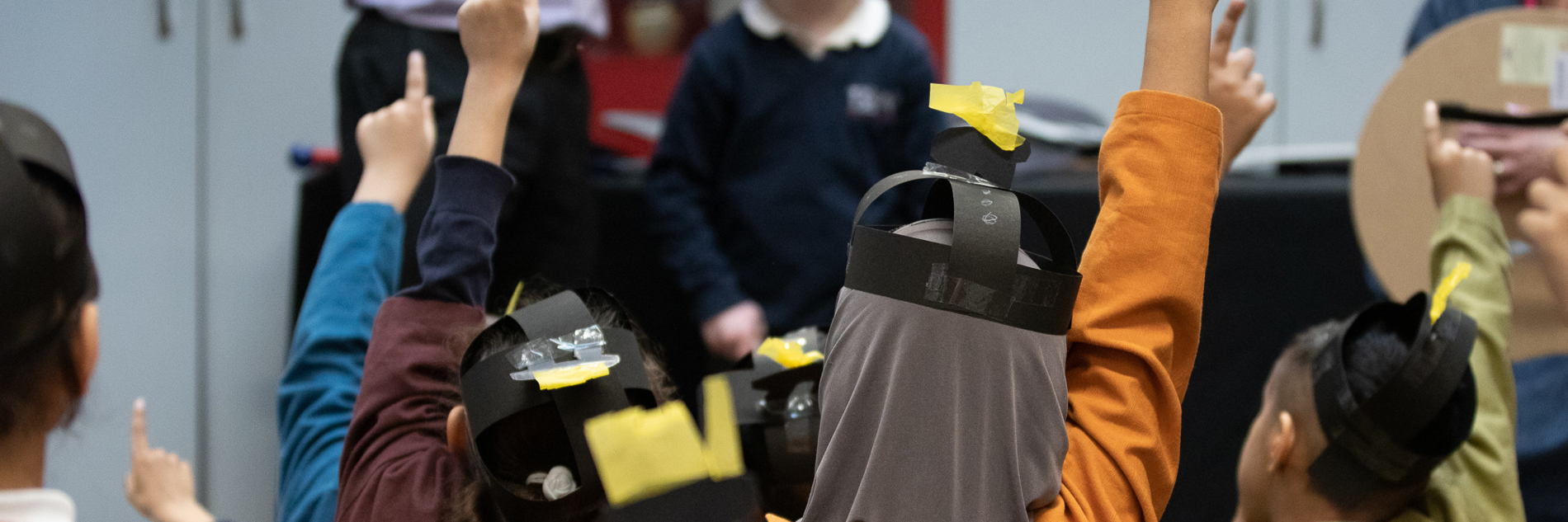 A view from behind of several primary school aged children in a school session, showing their heads, shoulders and raised hands. They are dressed as Anglo Saxons and wearing paper helmets.		

