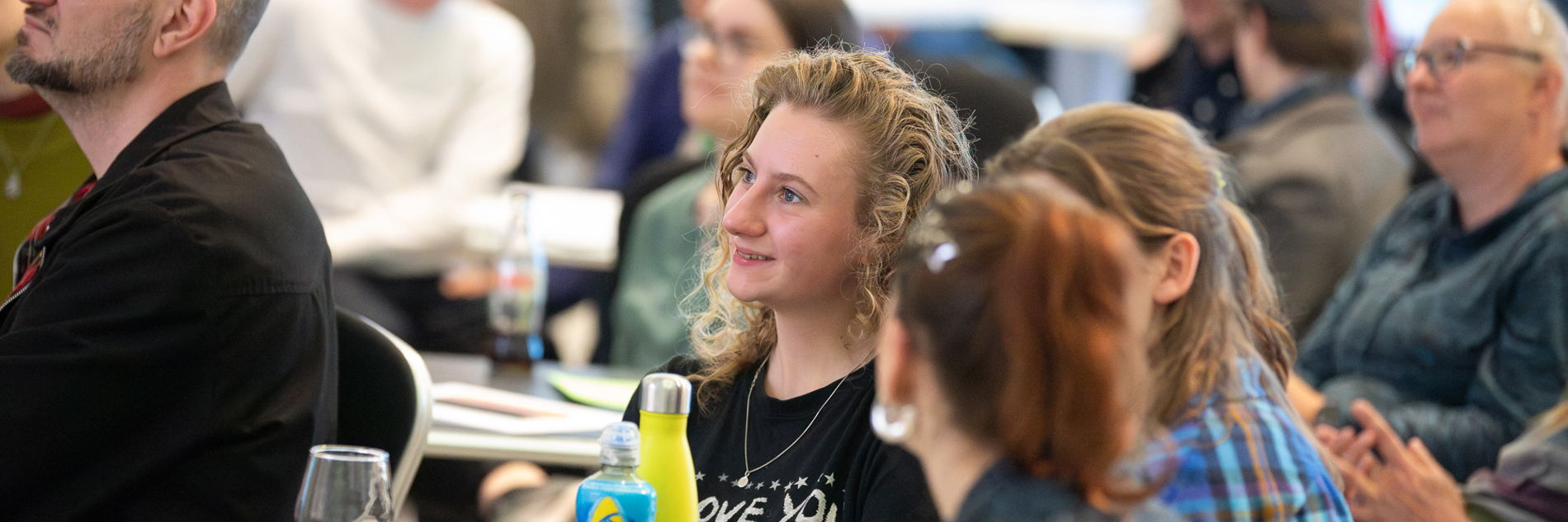 A photograph showing attendees at an event in Millennium Gallery. The room is full of people sat at tables, but the camera focuses on a person at the front with long curly blonde hair. On the table in front of them there is a glass or red wine, a bottle of lucozade and a bright yellow water bottle. There are also pencils, pens and paper on the table. Other people sit at the table and they seem to be in discussion