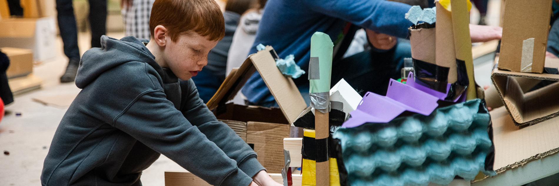 A child wearing a grey hoody and brown trousers kneeling on the floor works on a construction made of cardboard packaging and tubes, crates and gaffer tape. In the background adults and children work on other constructions.