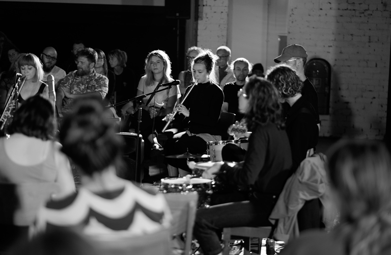A photograph of a group of adults playing musical instruments. An audience of adults are sitting on chairs surrounding the performers.  
