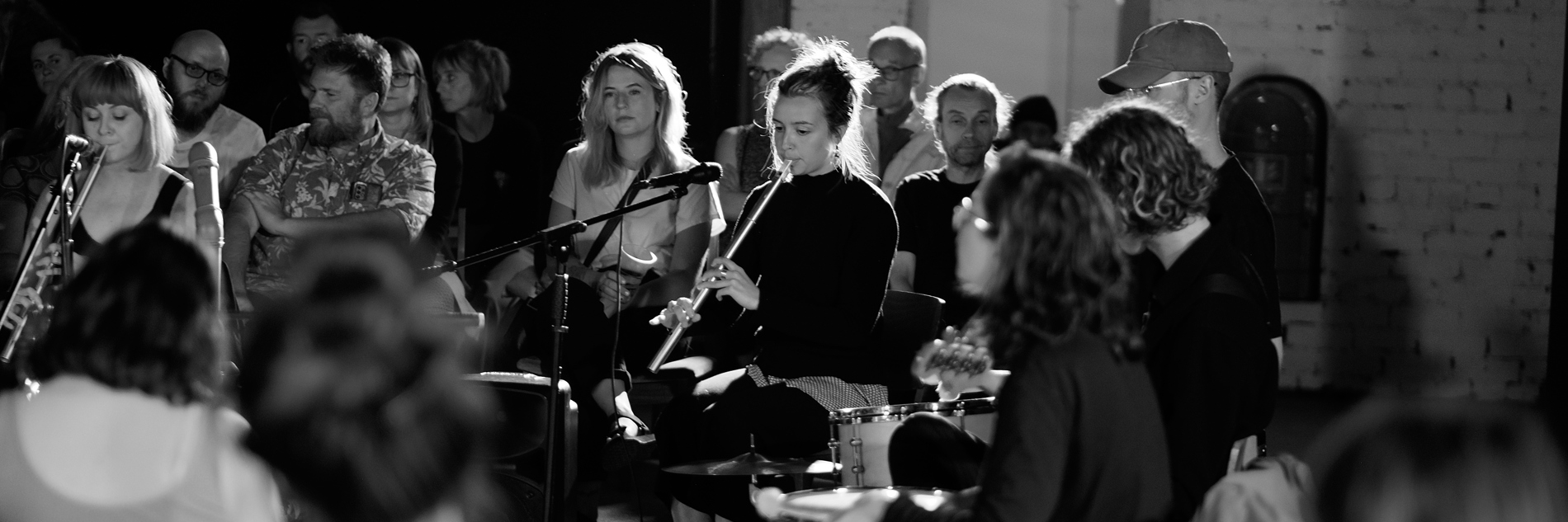 A photograph of a group of adults playing musical instruments. An audience of adults are sitting on chairs surrounding the performers.  