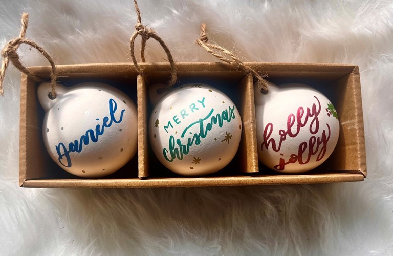 A photograph of three white baubles in brown cardboard packaging, photographed against a fluffy white background. The baubles all have calligraphy on them. From the left to right, they read 'Daniel' in blue, 'Merry Christmas' in green and 'Holly Jolly' in red.