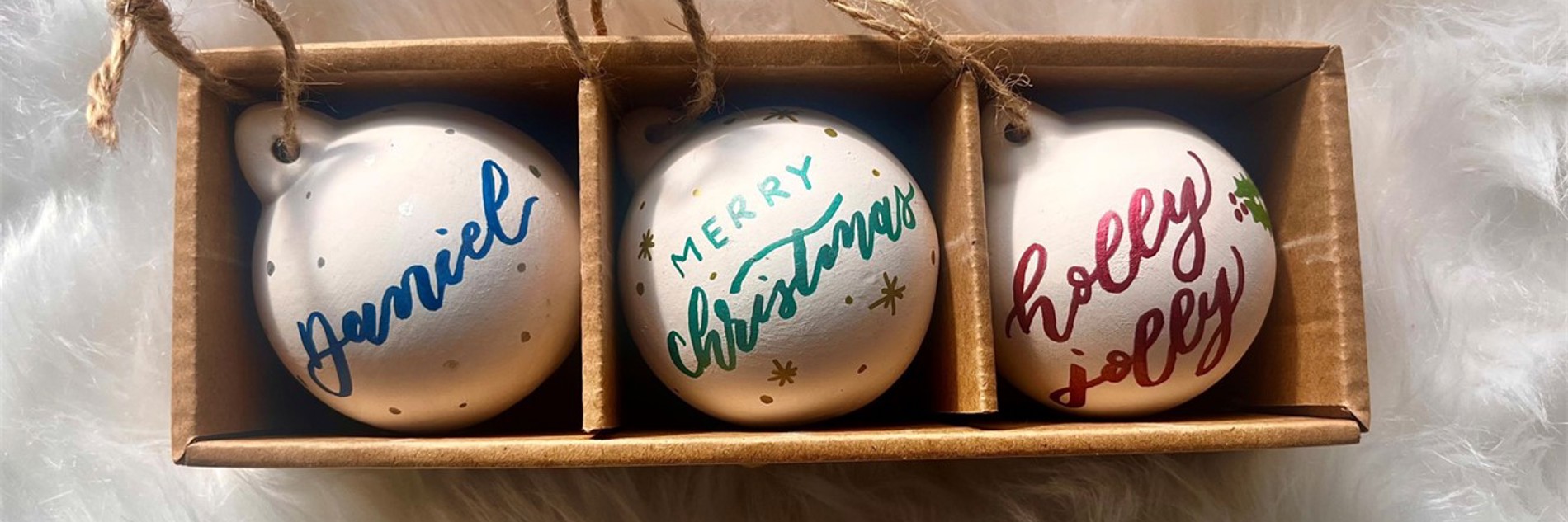 A photograph of three white baubles in brown cardboard packaging, photographed against a fluffy white background. The baubles all have calligraphy on them. From the left to right, they read 'Daniel' in blue, 'Merry Christmas' in green and 'Holly Jolly' in red.