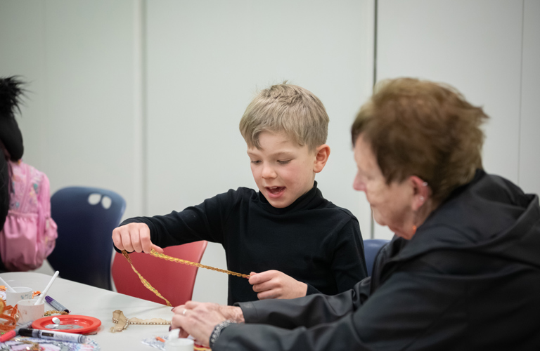 A photograph of a boy with blonde hair and wearing a black shirt holding a piece of decorative trim. An older lady sits beside him
