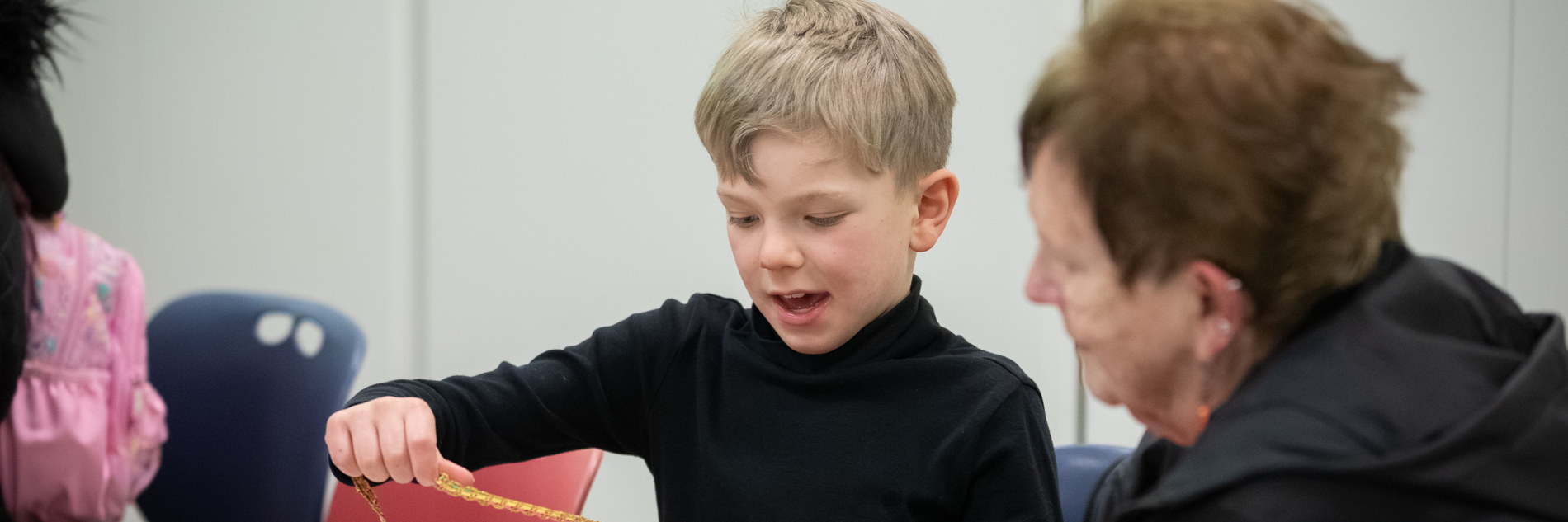 A photograph of a boy with blonde hair and wearing a black shirt holding a piece of decorative trim. An older lady sits beside him